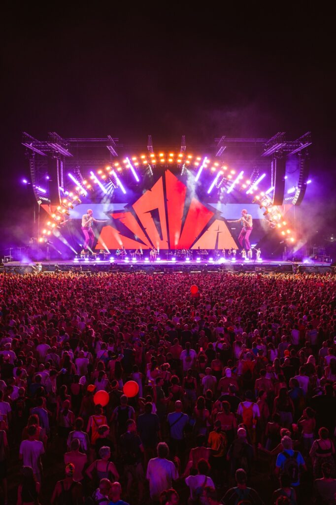 Panoramica del palco del Jova Summer Party con archi di luce viola, proiettori Chauvet e grande pubblico.